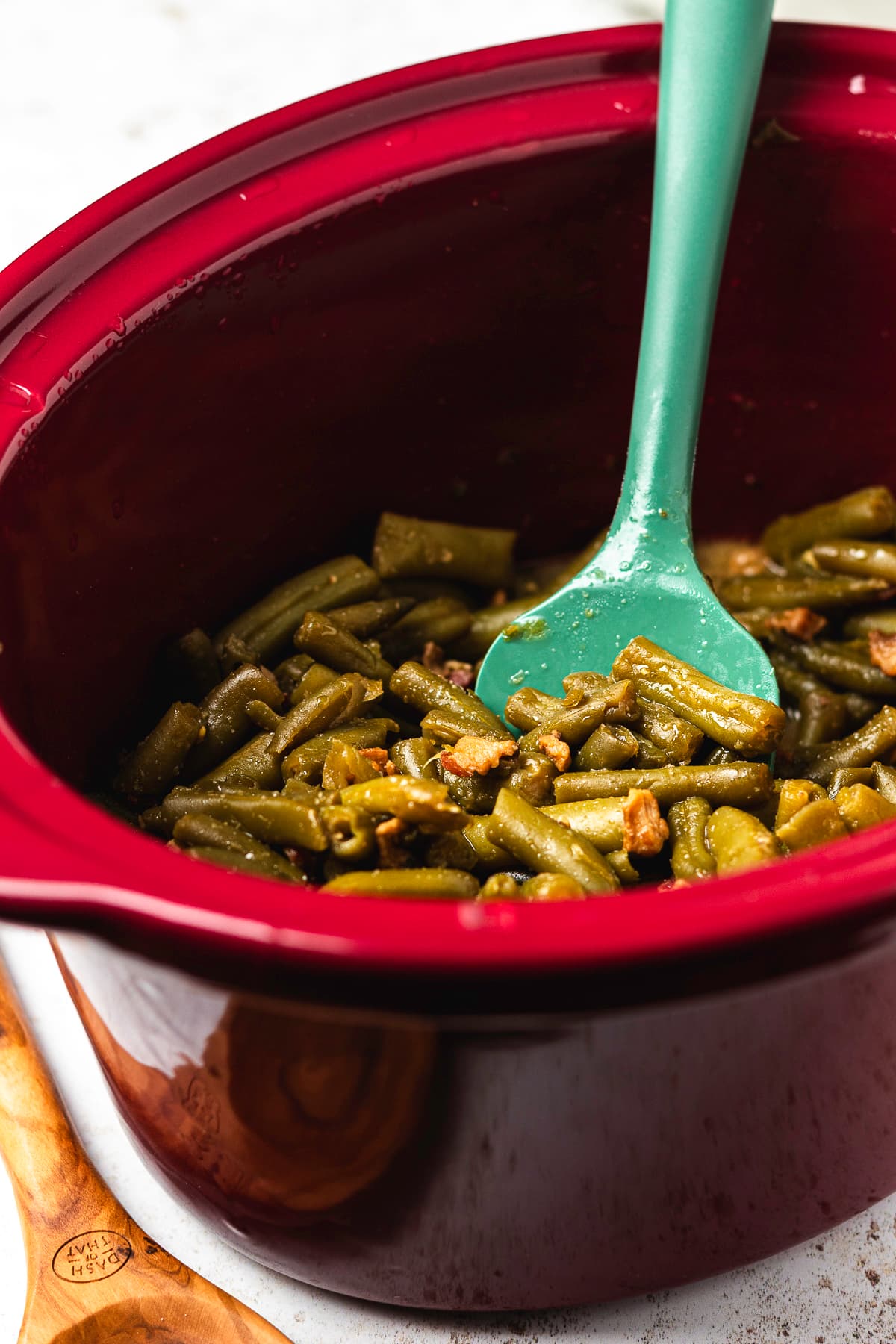 Finished crack green beans coated in brown sugar glaze and stirred with a green spatula in a red slow cooker.