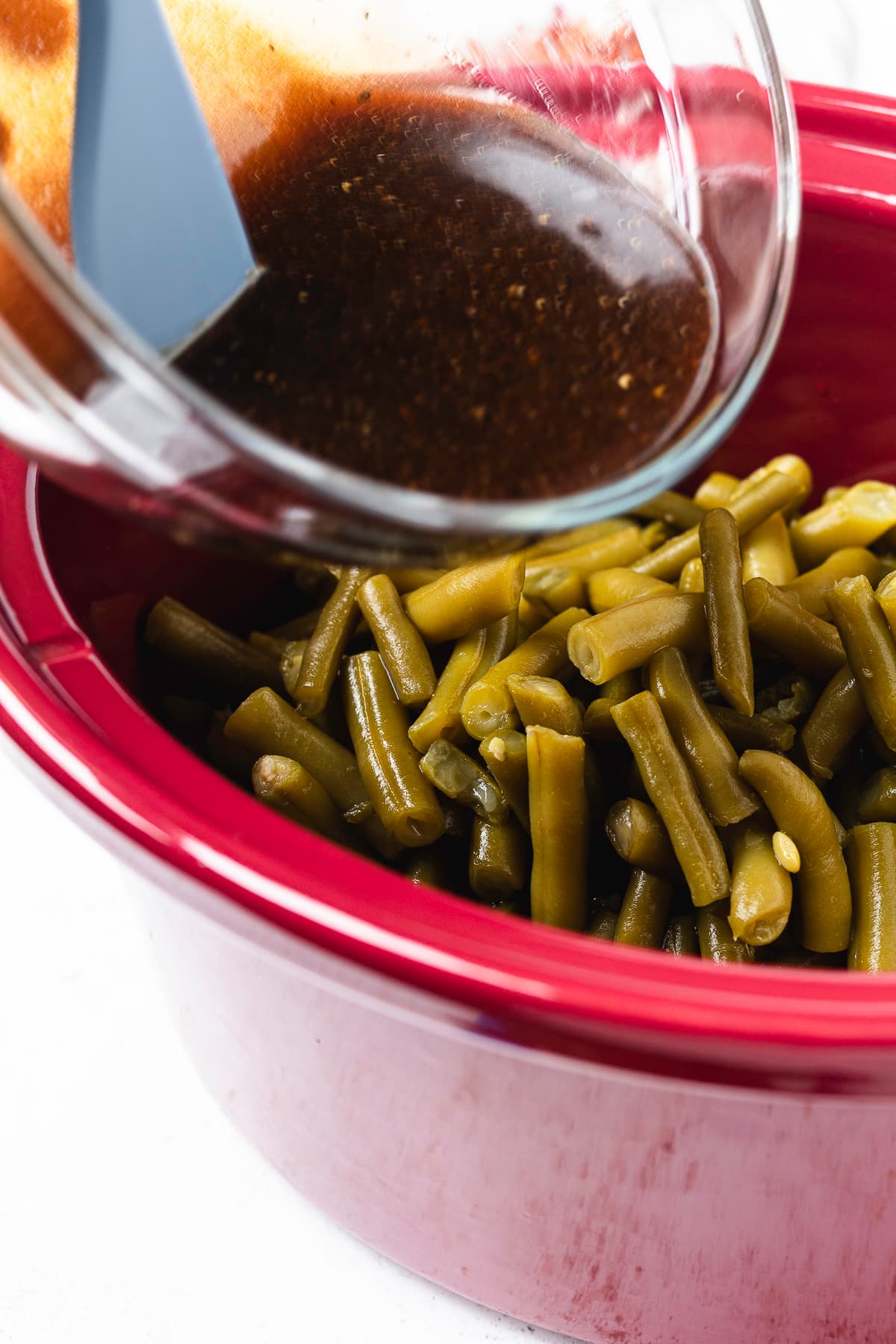 Pouring brown sugar sauce over green beans in a red slow cooker.