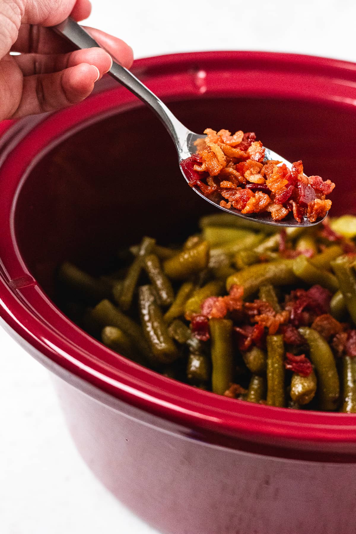 A spoonful of crispy chopped bacon being added to glazed green beans in a slow cooker.