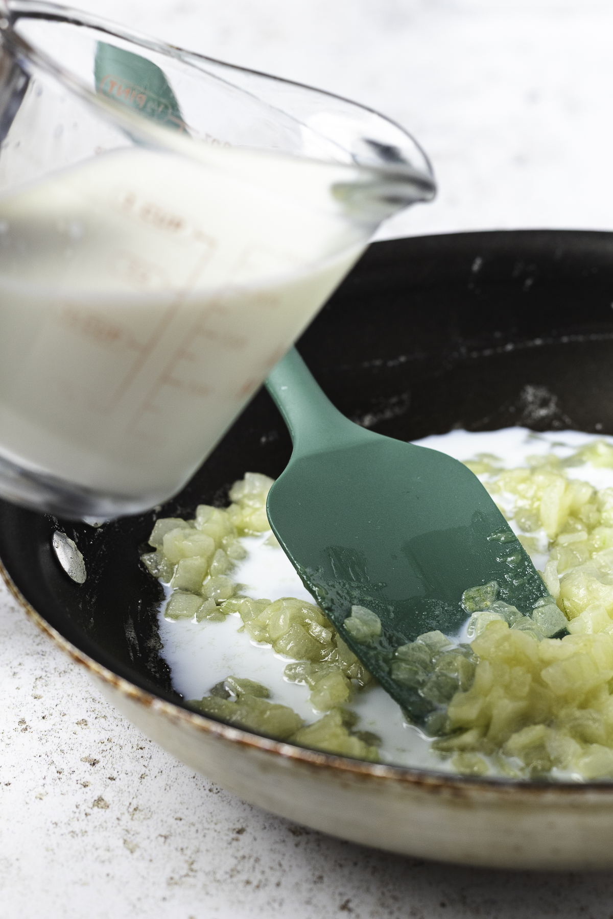 Milk being poured into a skillet with sautéed onions and butter to make stovetop creamed corn.