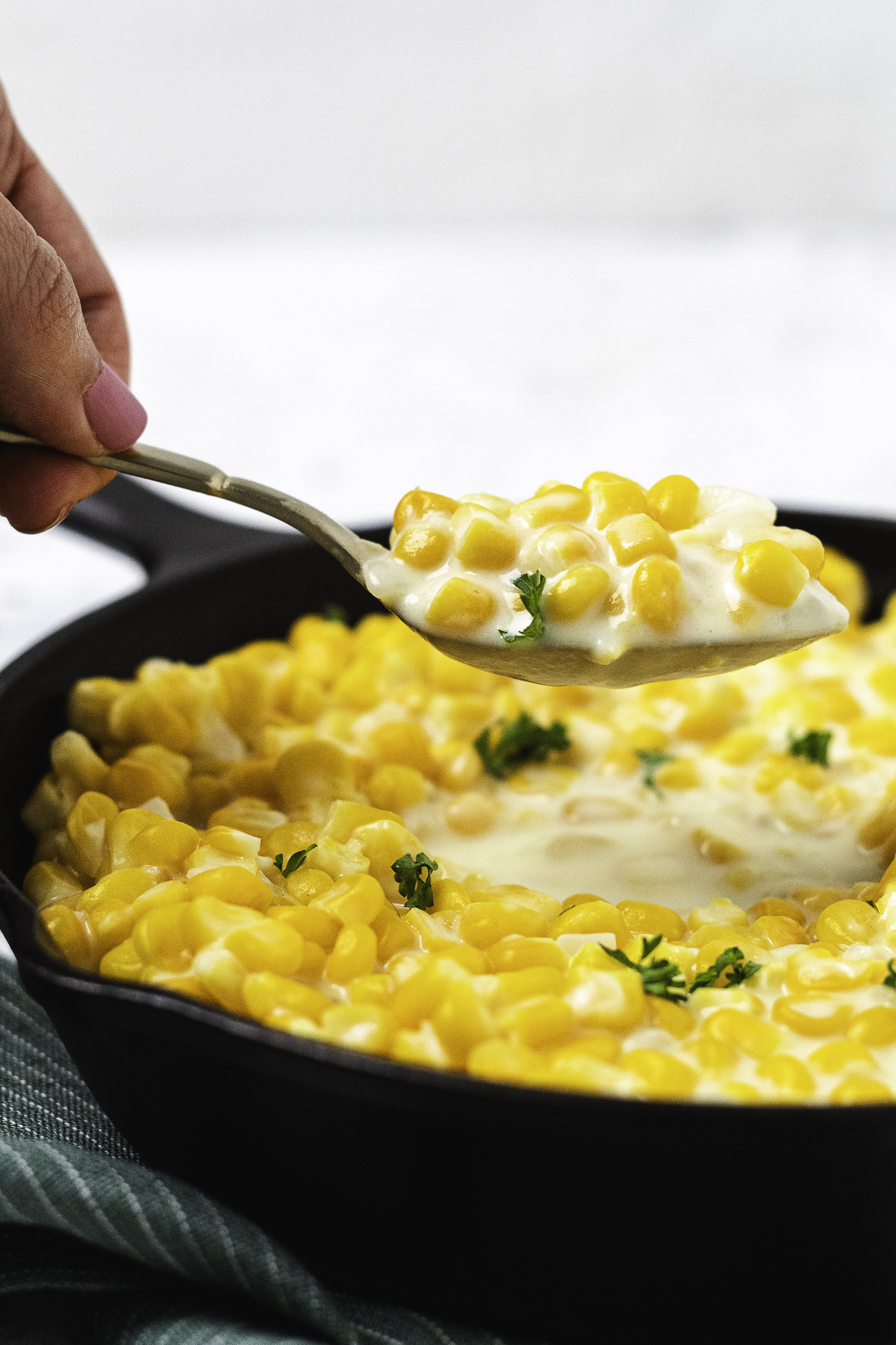 A spoonful of creamy sweet corn lifted from a black skillet garnished with fresh parsley.