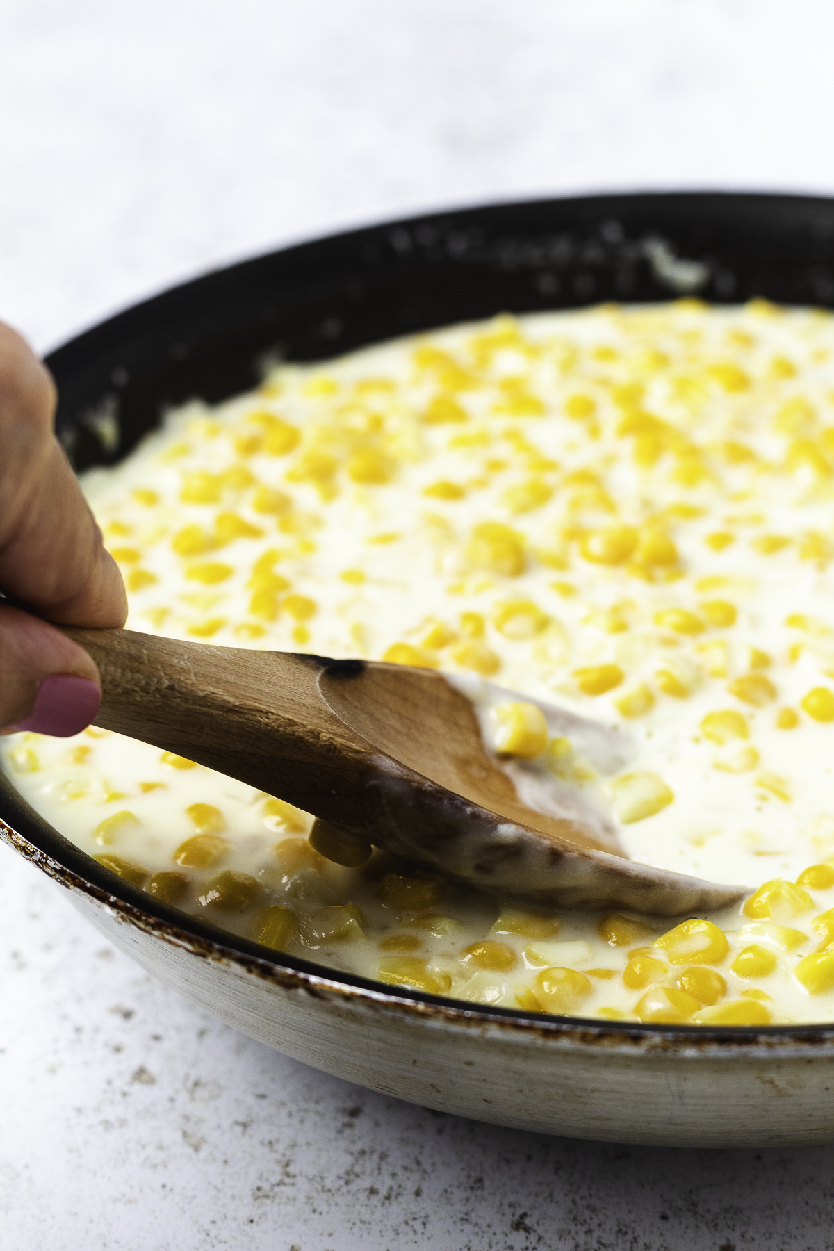 A hand stirs homemade stovetop creamed corn in a skillet with a wooden spoon.
