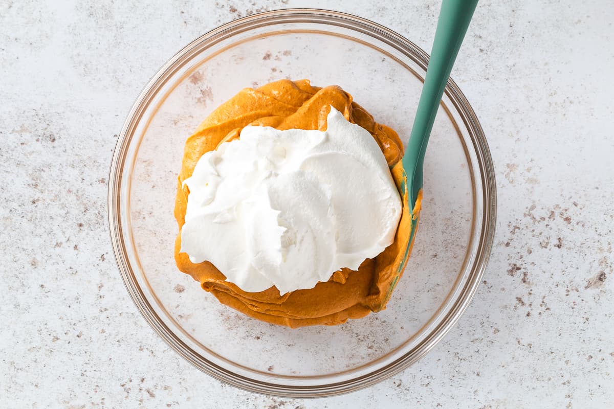 Pumpkin cheesecake mixture topped with whipped cream in a glass mixing bowl.